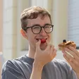 Cheerful young man with glasses biting into a strawberry held in a donut with a peanut butter topping, with a bright building in the background.