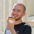 Smiling male person with short hair who holds a donut with chocolate icing and colorful sprinkles in front of him