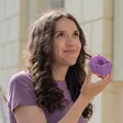 A woman with long curly brown hair, looking up thoughtfully, holding a purple sprinkle-covered doughnut, with sunlit windows in the background.