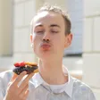 Young man playfully puckering his lips to kiss a strawberry atop a chocolate-frosted doughnut, with a bright building background.